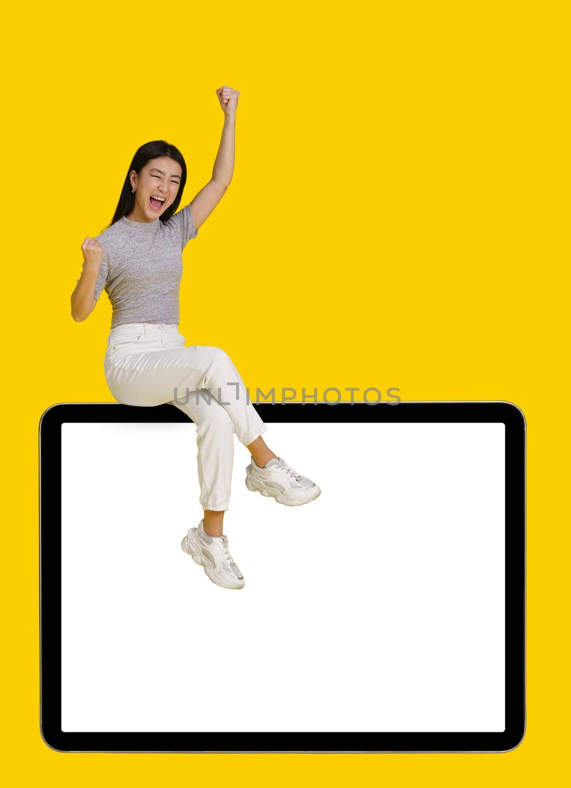 Win gesture excited asian girl sitting on giant, huge digital tablet pc with white blank screen isolated on yellow background. Mock up product placement for mobile app advertisement. Copy space by LipikStockMedia