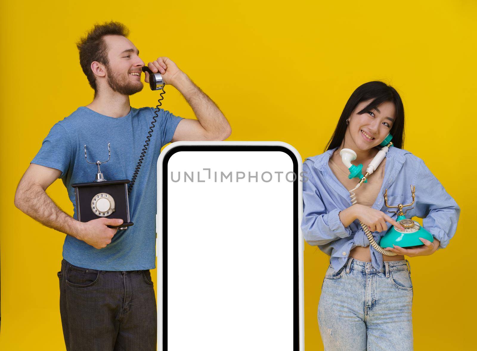 Handsome guy and asian girl talking on vintage phones leaned on huge smartphone or digital tablet with blank screen, happy smiling isolated on yellow background. Mock up, product placement.