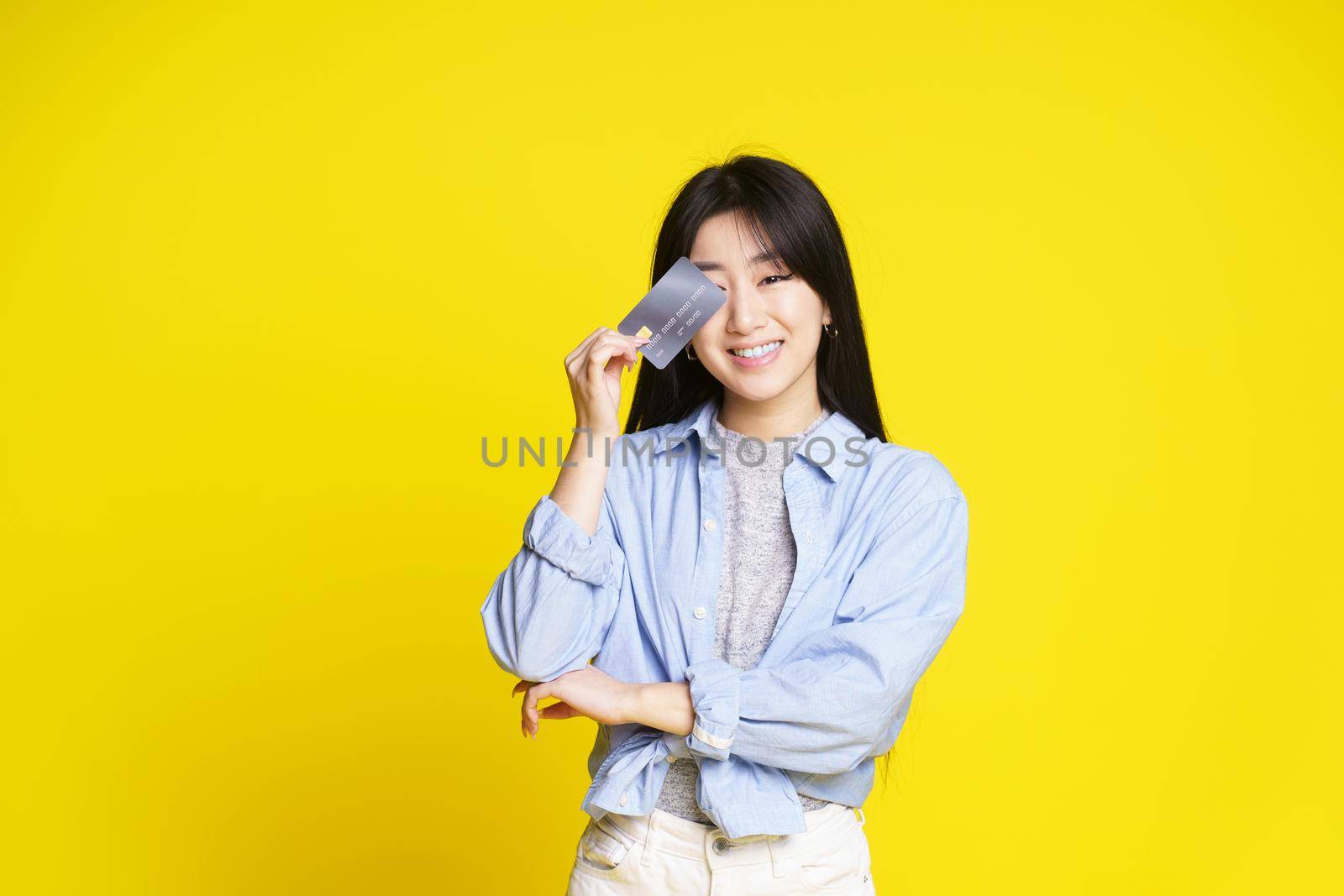 Charming asian girl holding credit, debit card touching her eye. Asian girl in blue shirt holding mockup credit card isolated on yellow background. Shopping concept. Banking concept by LipikStockMedia