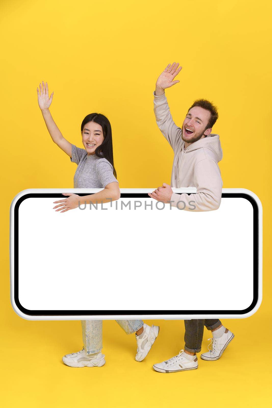 Greeting handsome guy and asian girl holding huge smartphone, digital tablet with white screen, mobile app advertisement and excited smile on camera isolated on yellow background. Product placement.