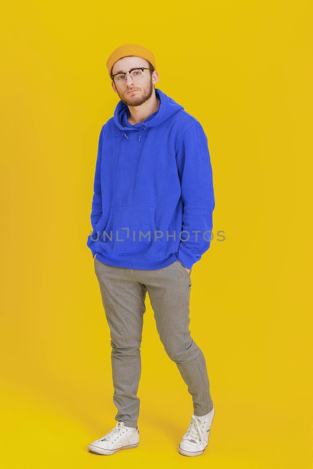 Young man model in casual wear, glasses posing looking at camera on yellow background with hands in pockets. Stylish bearded smart hipster man casual look. Thoughtful guy on yellow background.