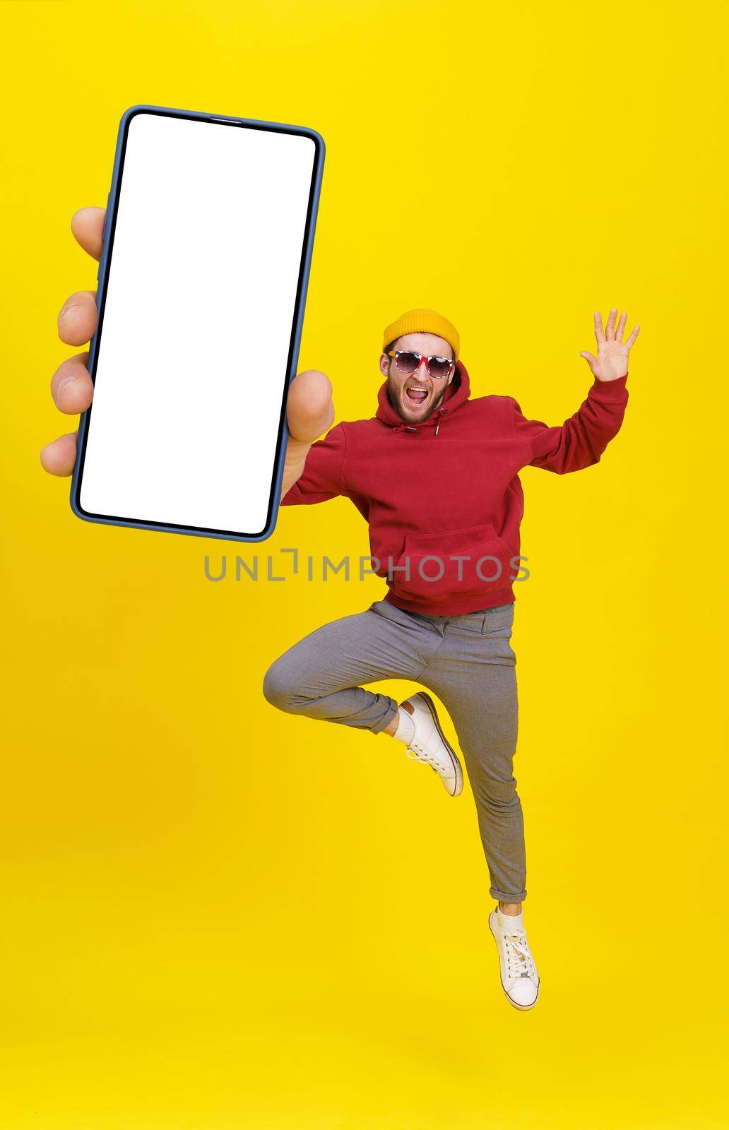 Young man in Uk flag sunglasses jumping in joy like ballet dancer holding smartphone wearing casual red hoodie and jeans isolated on yellow background. Mobile app advertising product placement.