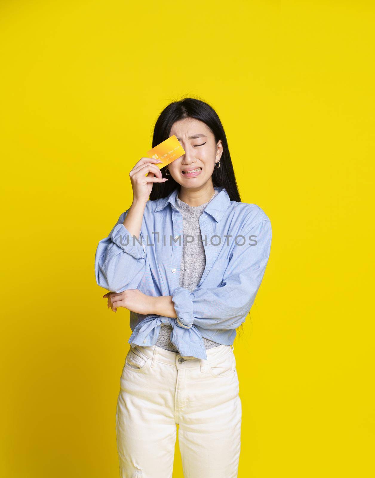 Unhappy, broke crying asian girl holding yellow credit, debit card next to her face begging for money. Asian girl in blue shirt with mockup credit card isolated on yellow background. Shopping concept by LipikStockMedia