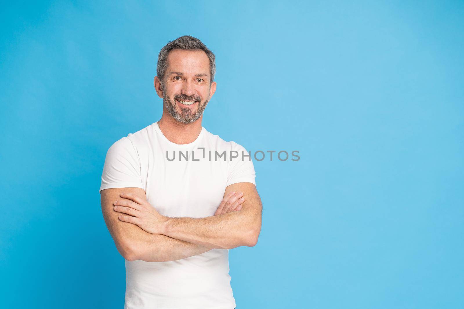 Middle aged grey haired man with standing with arms folded happy smiling on camera wearing white t-shirt isolated on blue background. Mature fit man, healthy lifestyle concept.