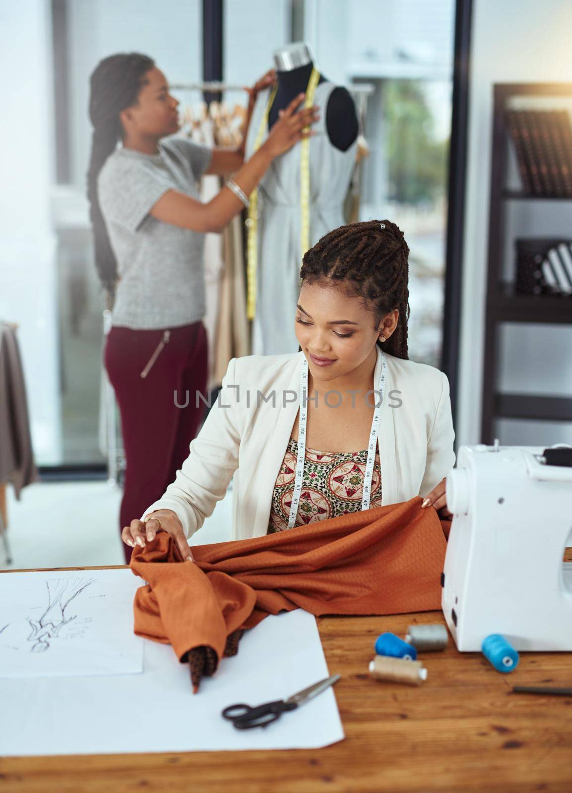 Shot of a young fashion designer sewing garments while a colleague works on a mannequin in the background.