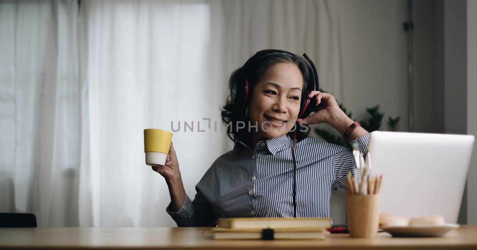 Happy senior asian woman wearing headphones listens songs on laptop in living room at cozy home by wichayada