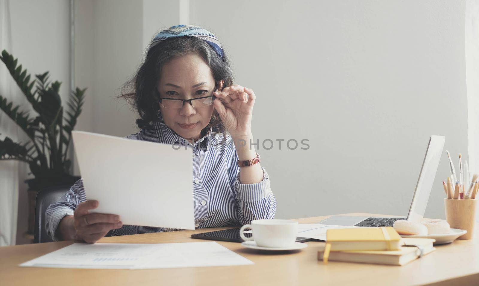 Seriously senior woman tworking at home, looking at laptop and waving paper correspondence, sitting at home, free space. by wichayada