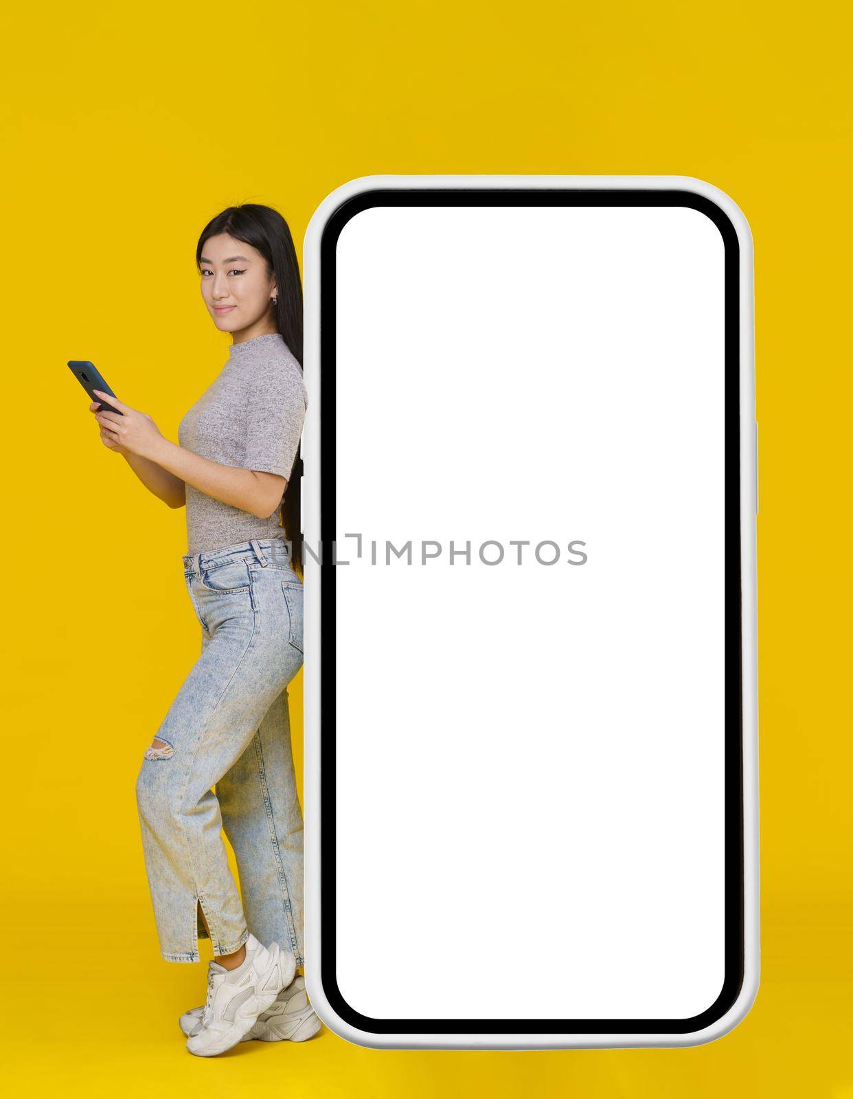 Smiling asian girl with phone in hands leaned back on giant, huge smartphone, digital tablet with white, black screen standing wearing casual isolated on yellow background. Free space mock up by LipikStockMedia