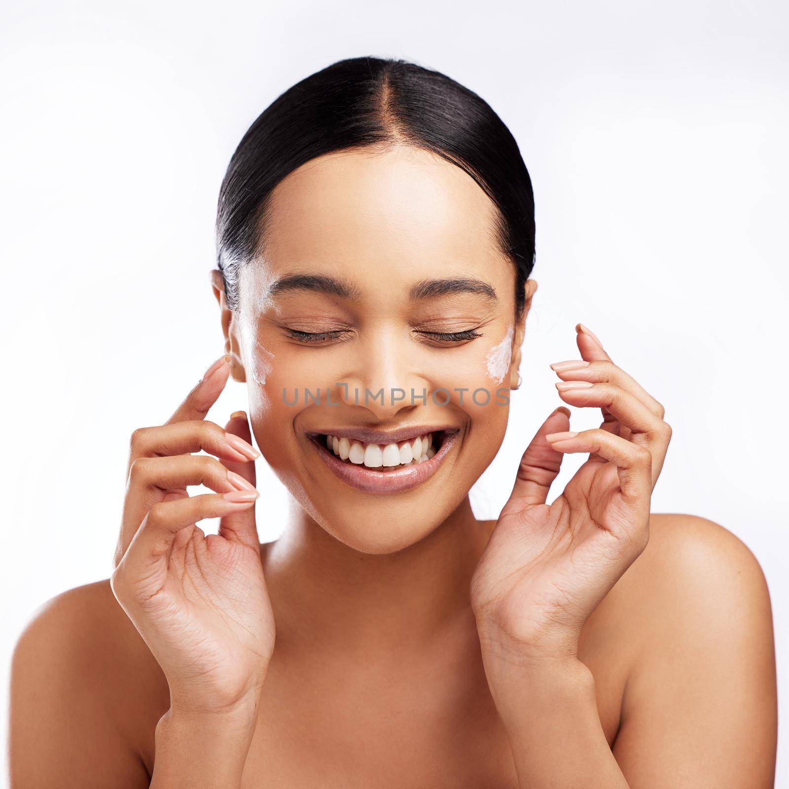 Studio shot of a beautiful young woman applying moisturiser to her face against a white background.