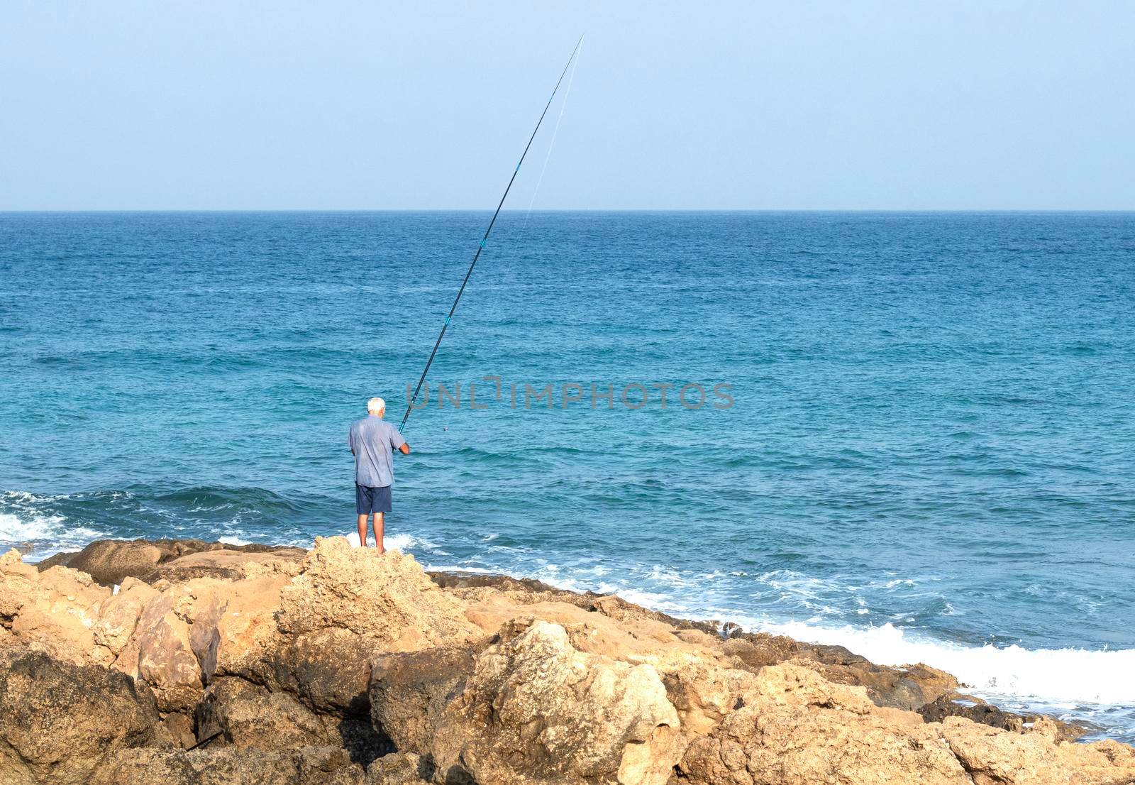 An unrecognizable male fisherman stands on the beach and throws a fishing rod. Sunny day. A lone fisherman tries his luck as the waves splash over the basalt rocks . High quality photo