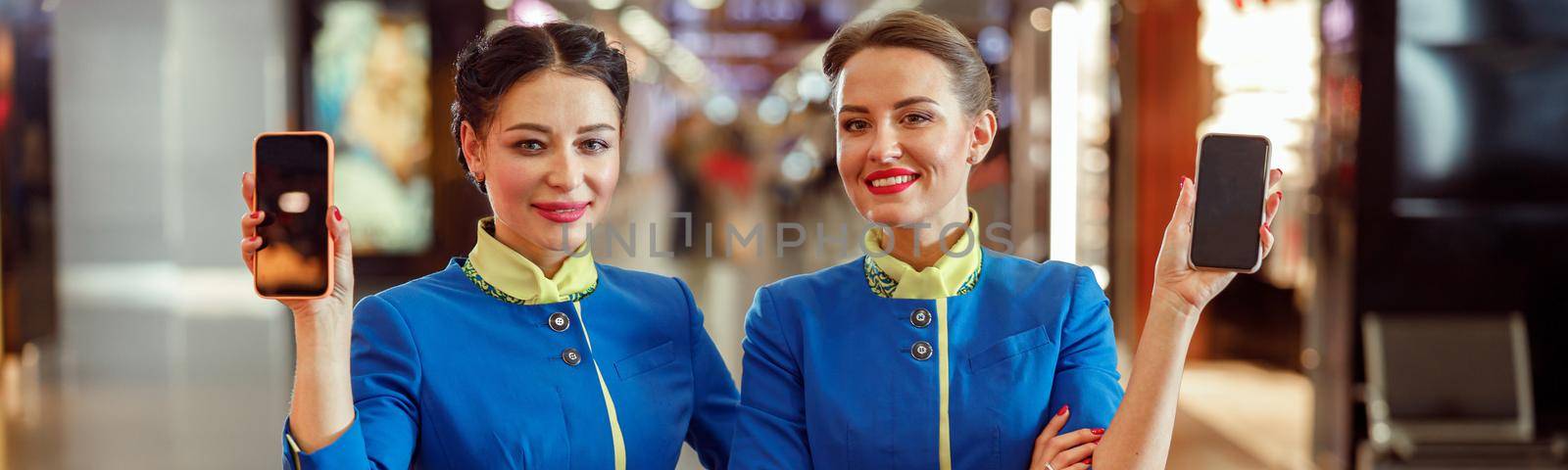 Joyful female flight attendants in aviation air hostess uniform looking at camera and smiling while holding mobile phones