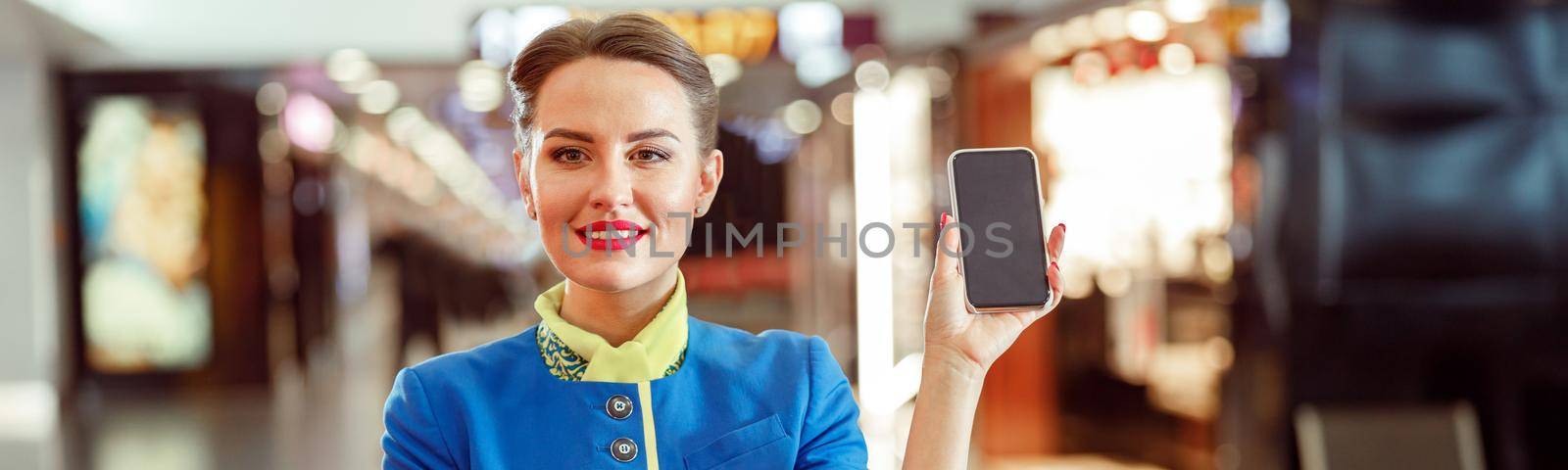 Joyful woman flight attendant in air hostess uniform looking at camera and smiling while holding modern smartphone