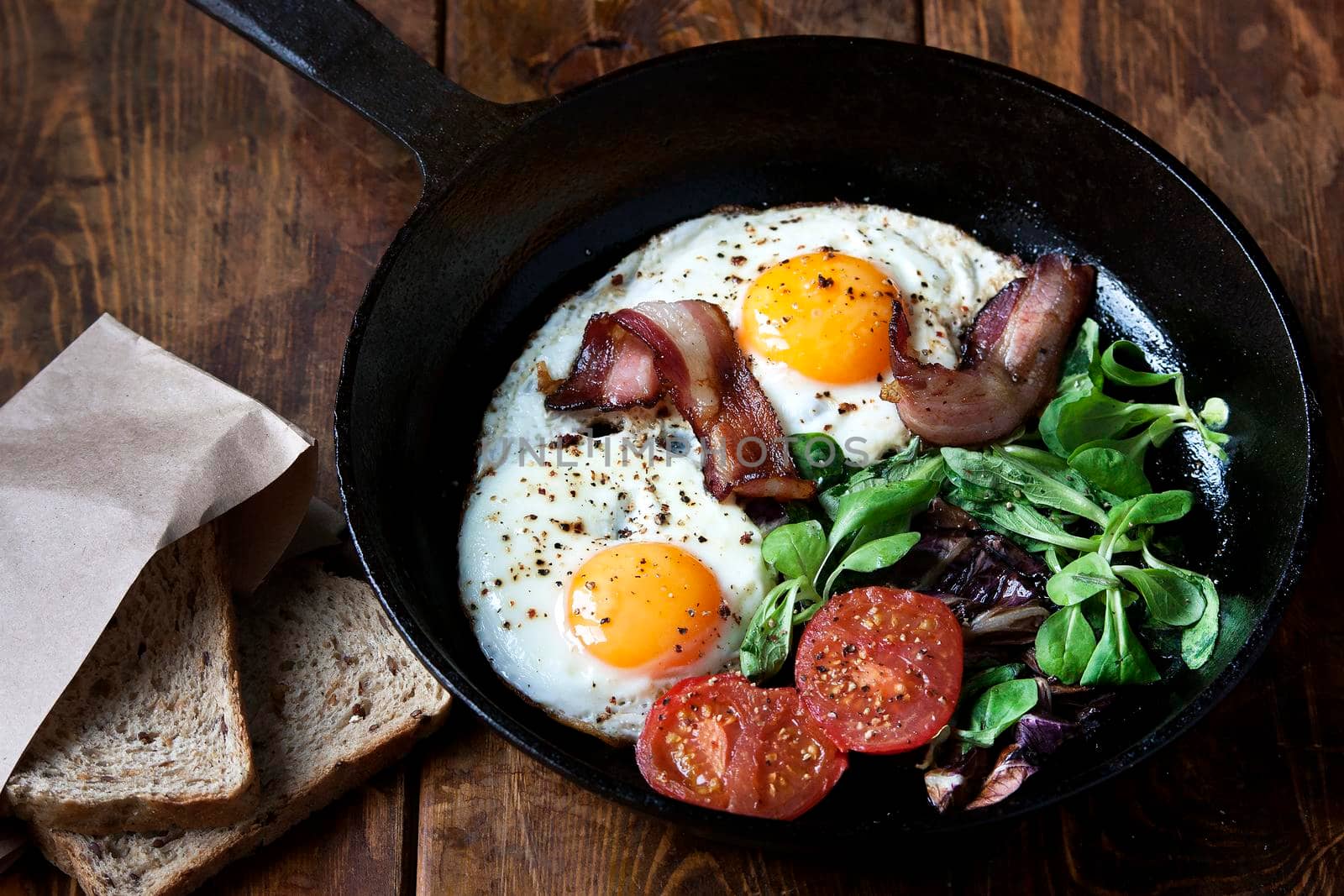 Breakfast set. Pan of fried eggs with bacon, fresh tomato, sage and bread on dark serving board over black wooden background, top view, copy space