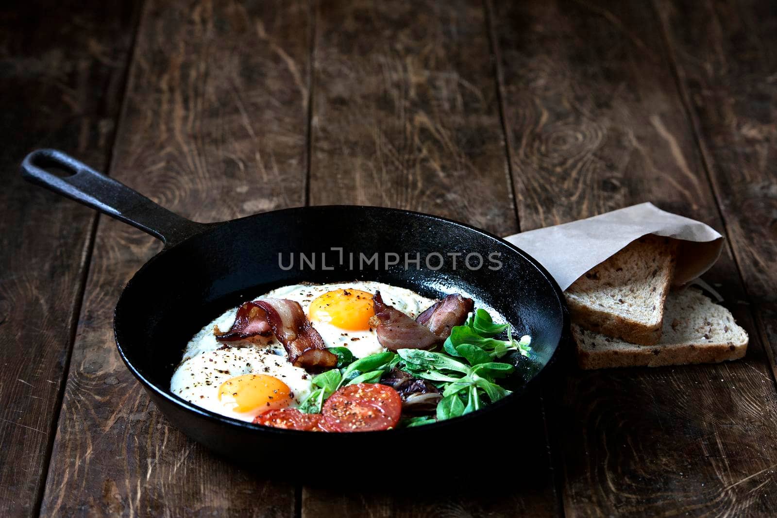 Breakfast set. Pan of fried eggs with bacon, fresh tomato, sage and bread on dark serving board over black wooden background, top view, copy space