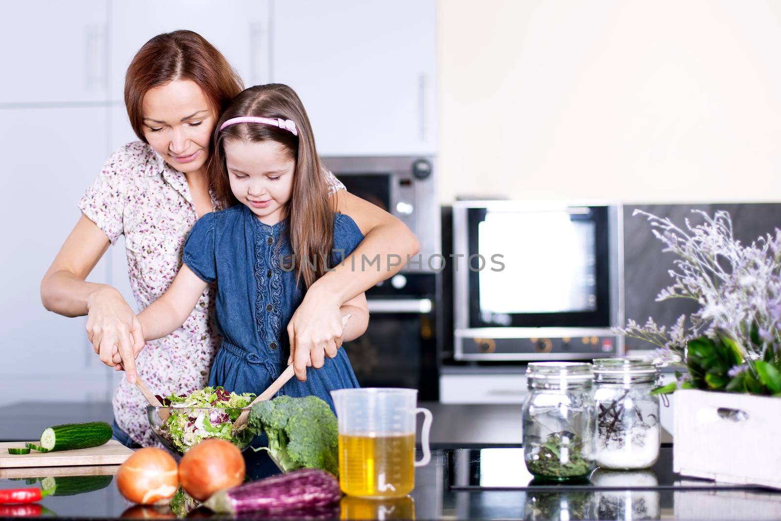 Mother with little daughter cooking in the kitchen at home. Girl Assisting In Preparing Food - Stock image