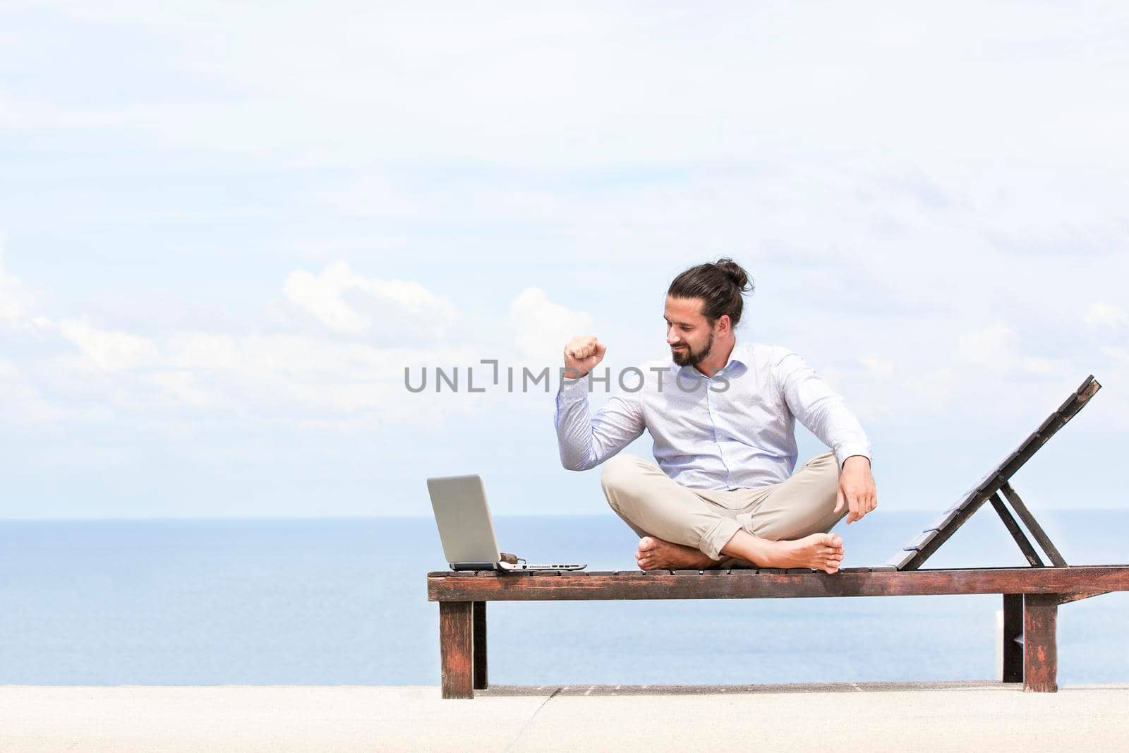 Young businessman on the beach resting on his deck chair using his tablet by Jyliana