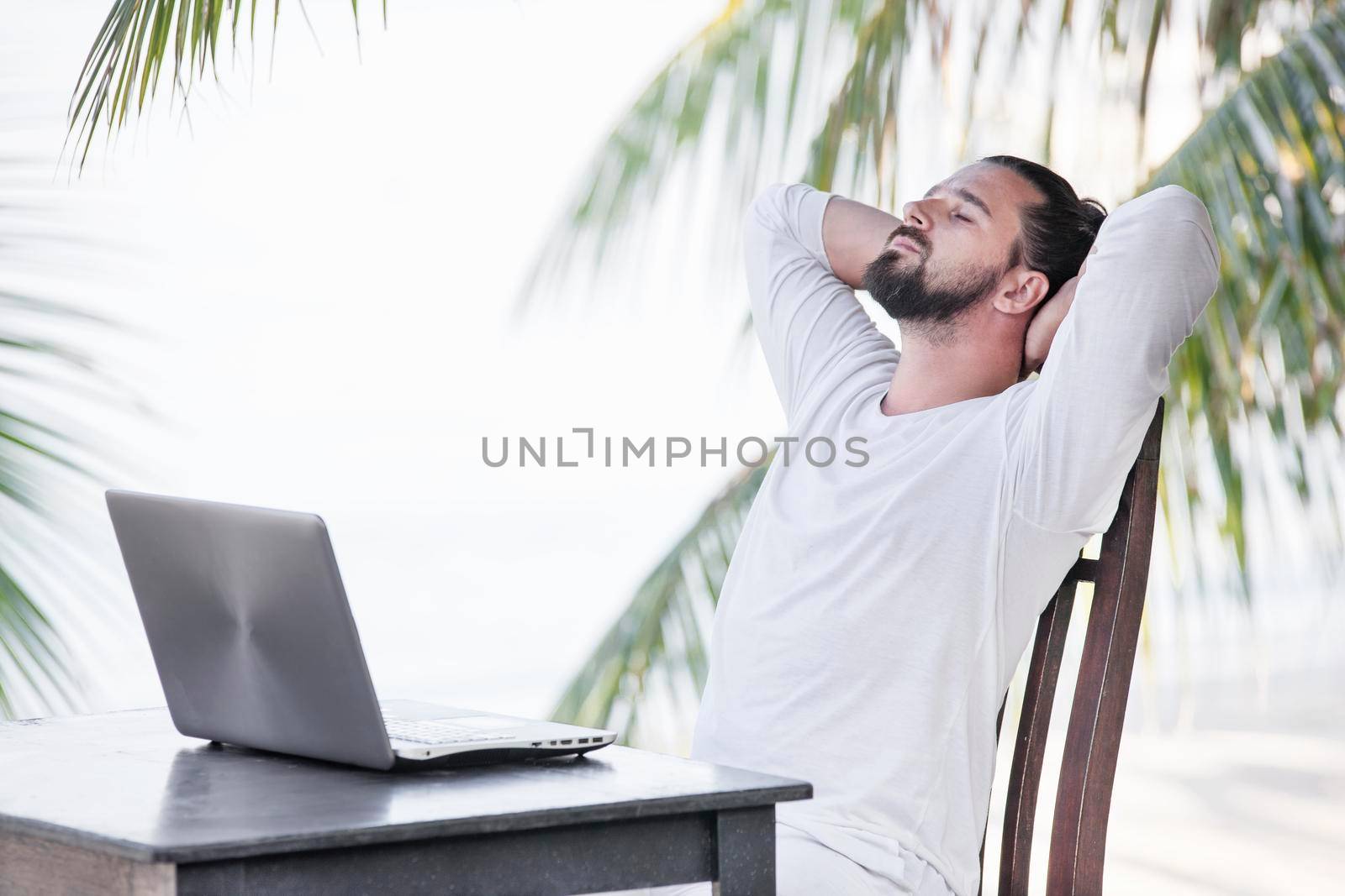 Vacation and technology. Work and travel. Young bearded man using laptop computer while sitting at beach cafe bar. by Jyliana