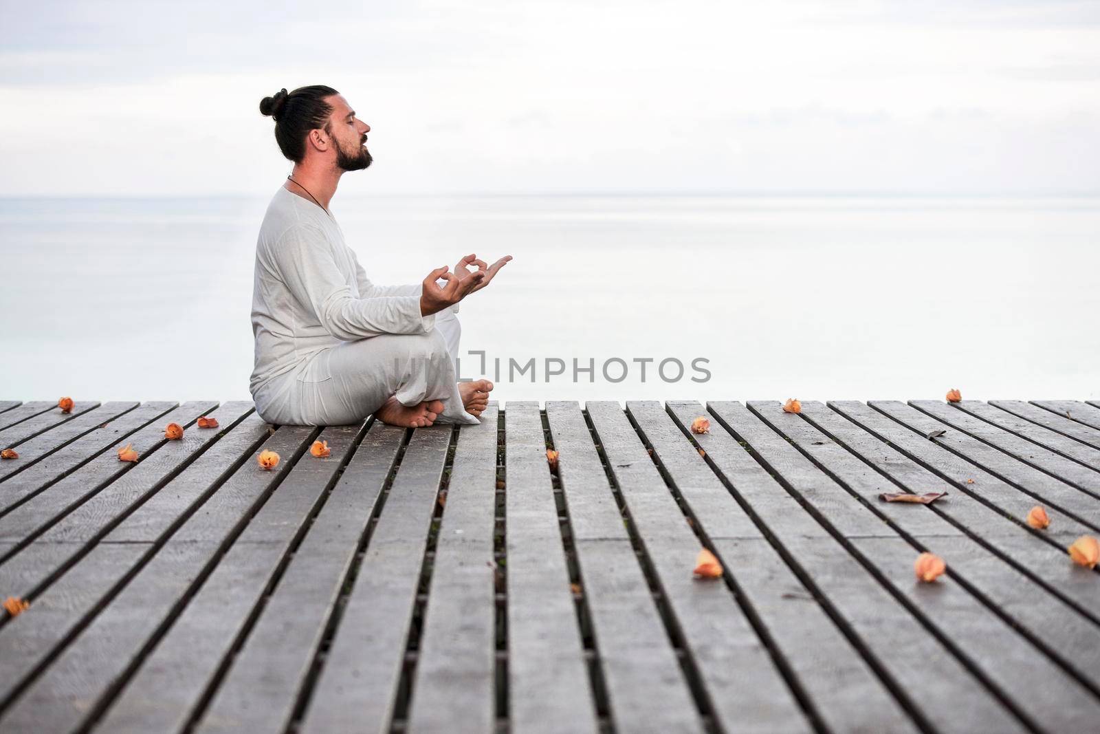 Man in white clothes meditating yoga on wooden pier near ocean