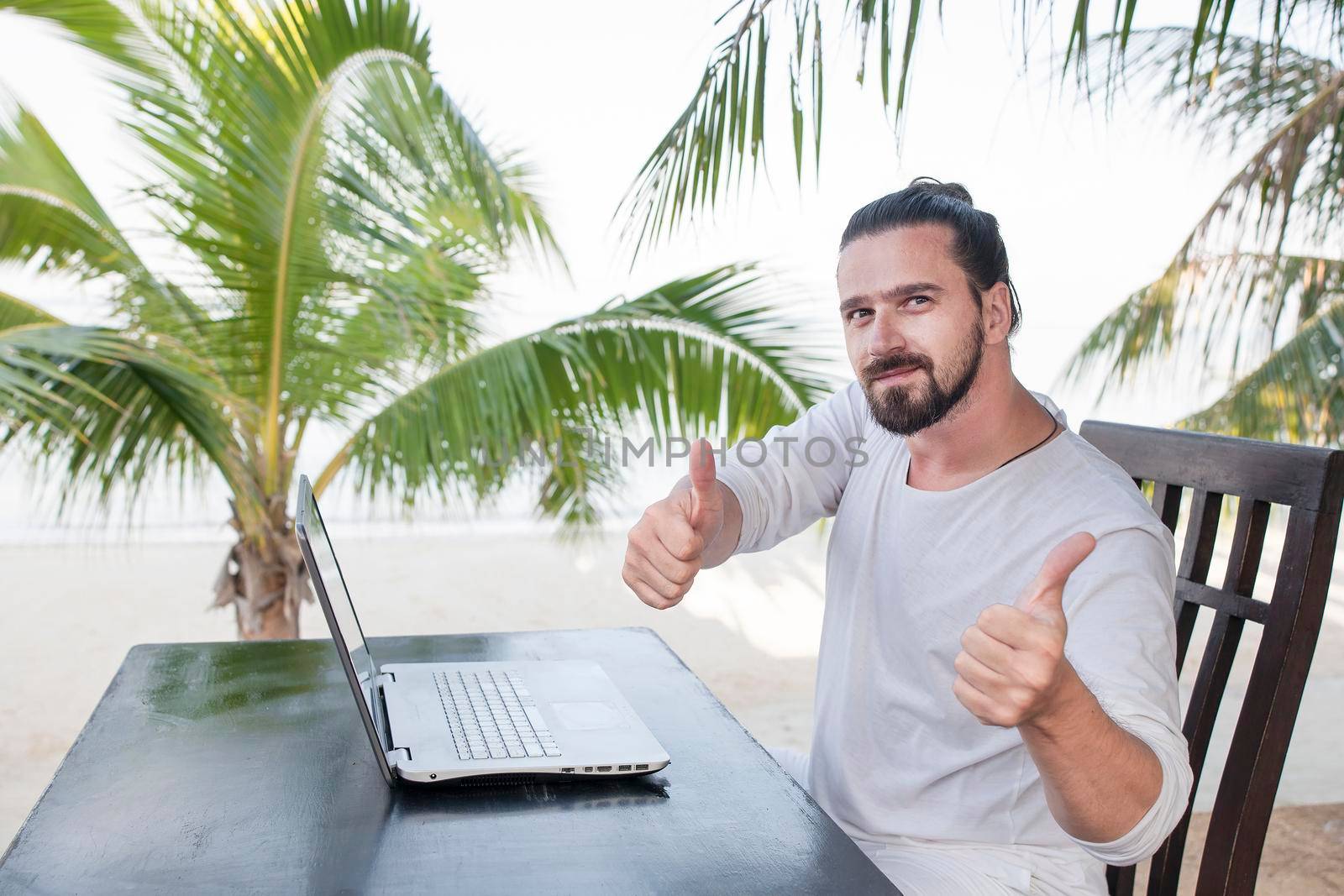 Vacation and technology. Work and travel. Young bearded man using laptop computer while sitting at beach cafe bar near palms and showing thumbs up