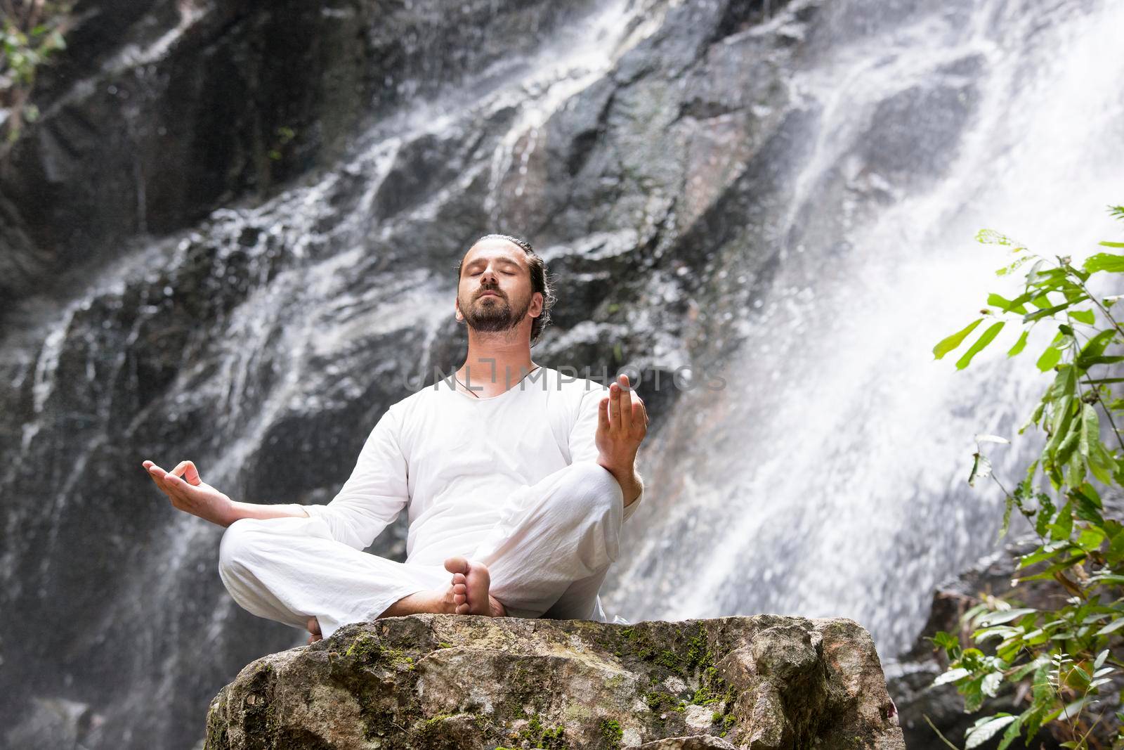 Wellness spa, vacation and yoga meditation concept. Young man sitting in lotus position on the rock under tropical waterfall.