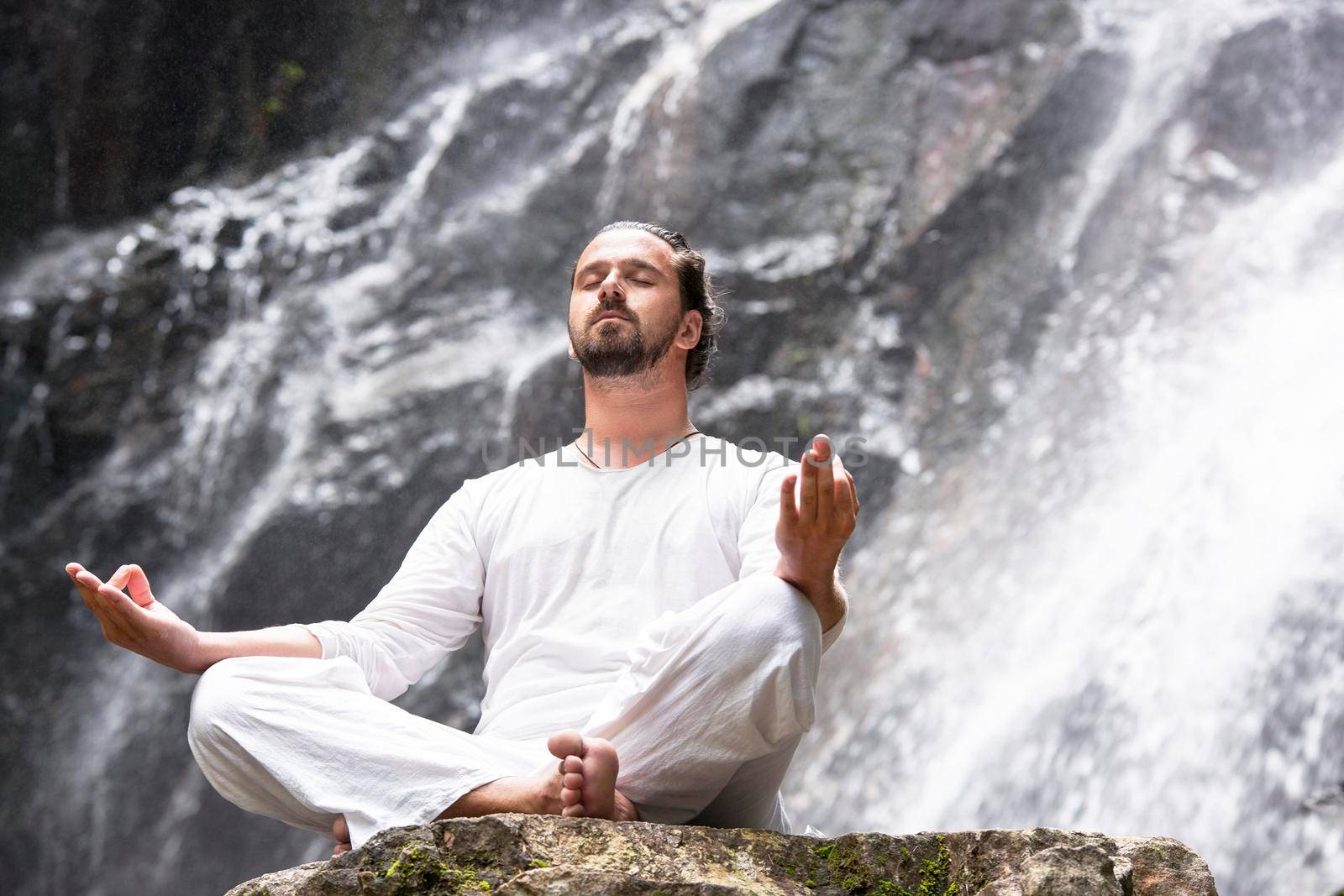 Wellness yoga meditation concept. Young man sitting in lotus position on the rock under tropical waterfall. by Jyliana