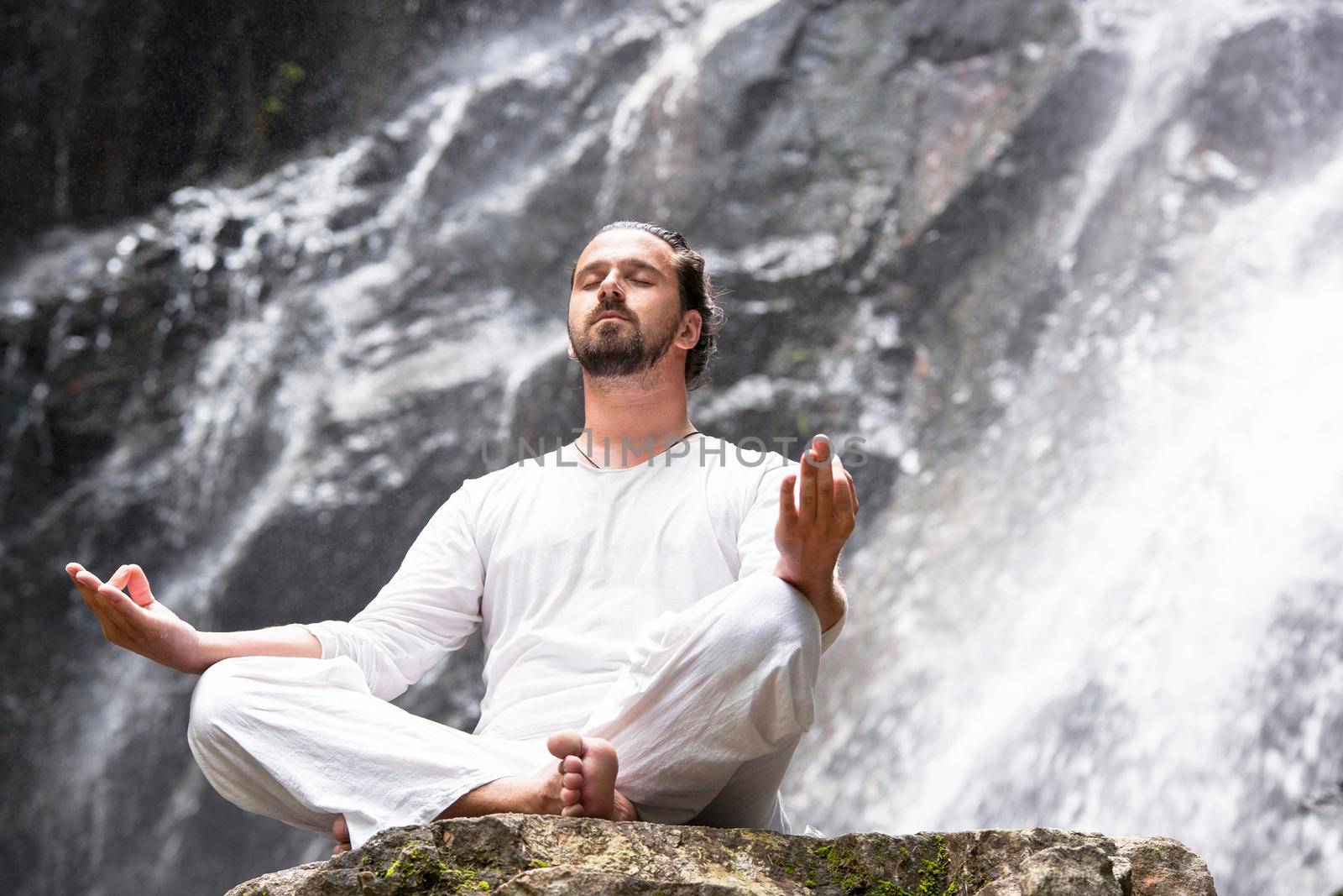 Wellness spa, vacation and yoga meditation concept. Young man sitting in lotus position on the rock under tropical waterfall.