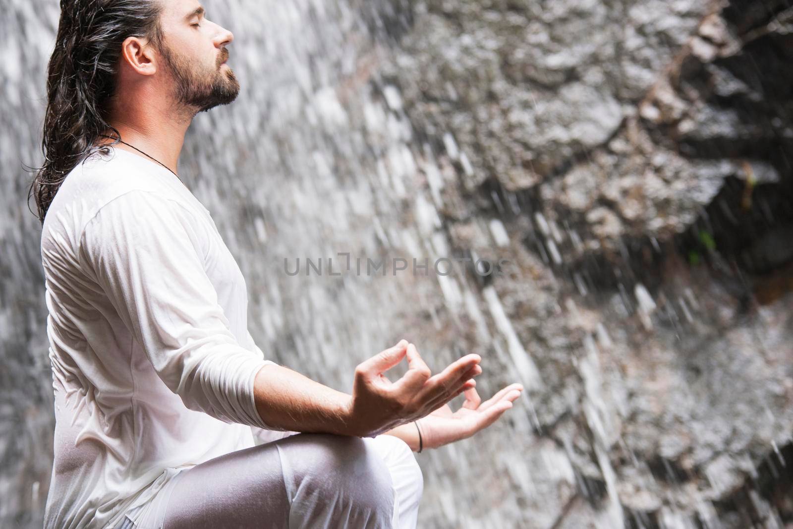 Wellness yoga meditation concept. Young man sitting in lotus position on the rock under tropical waterfall. by Jyliana