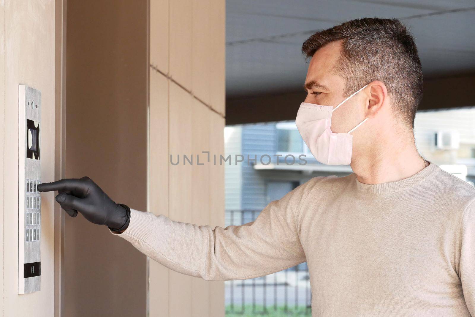 Man in face mask and gloves dials the code on the intercom and going in porch of his apartment. Pandemic covid-19 concept by Jyliana