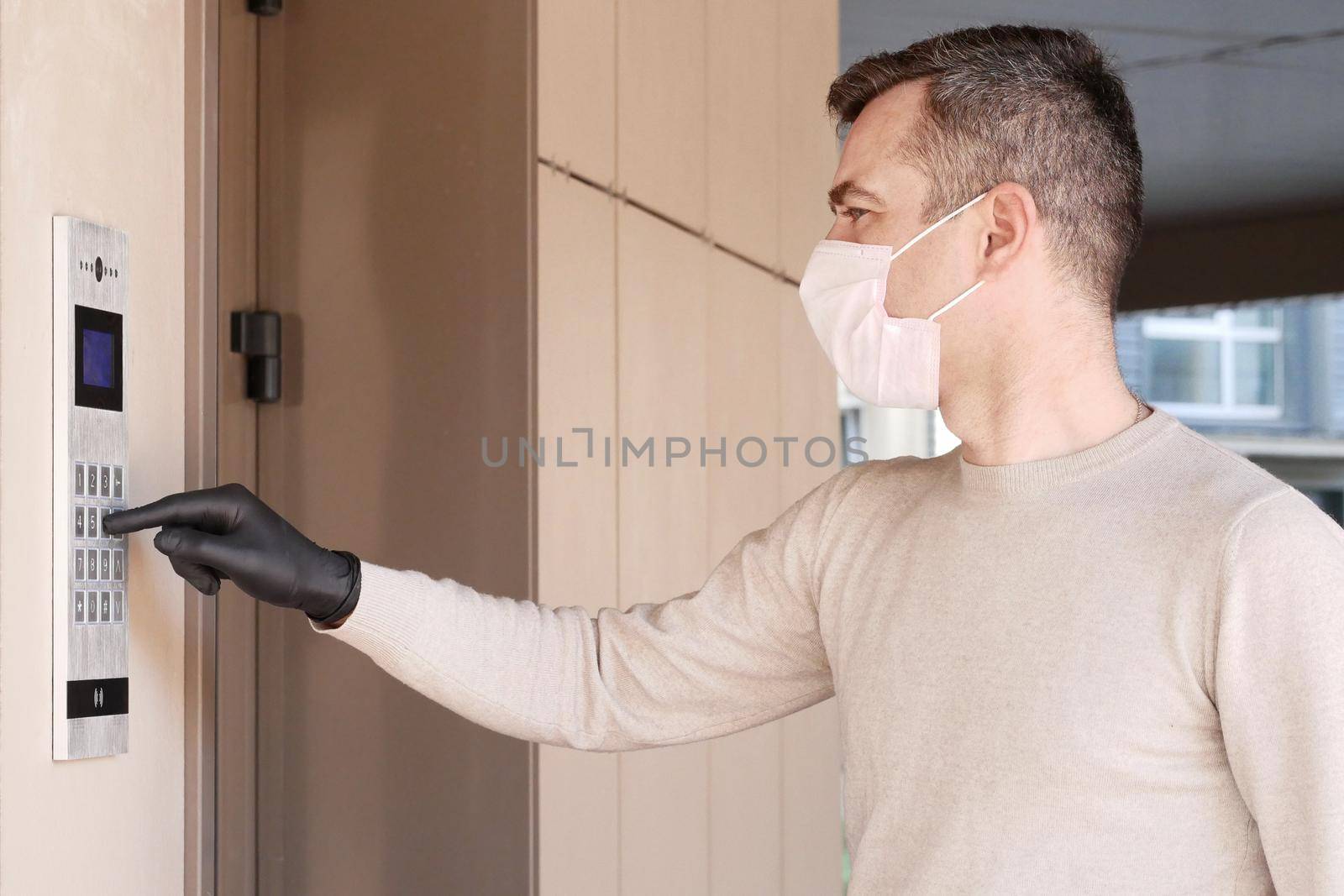 Man in face mask and gloves dials the code on the intercom and going in porch of his apartment. Pandemic covid-19 concept by Jyliana