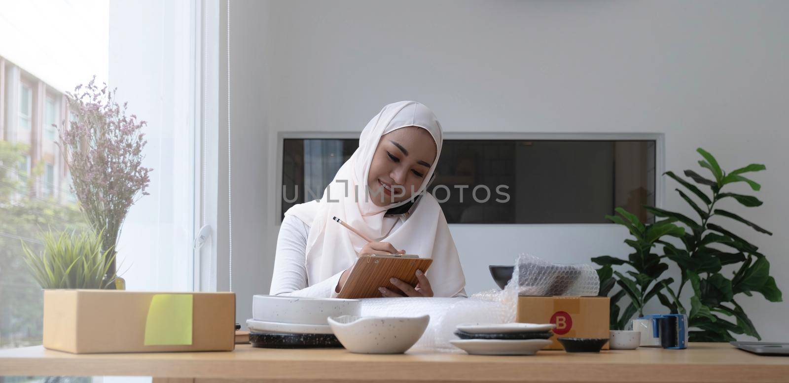 Religious asian muslim woman sitting and using smart mobile phone and talking to customer with happy face and business package SME box delivery background..