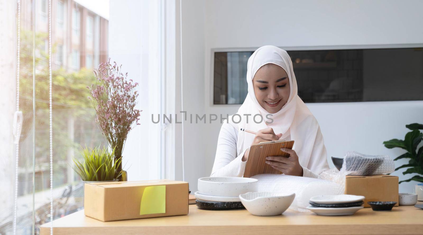 Professional and successful young muslim female small business owner talking on the phone with her client and noting an order on notepad. by wichayada