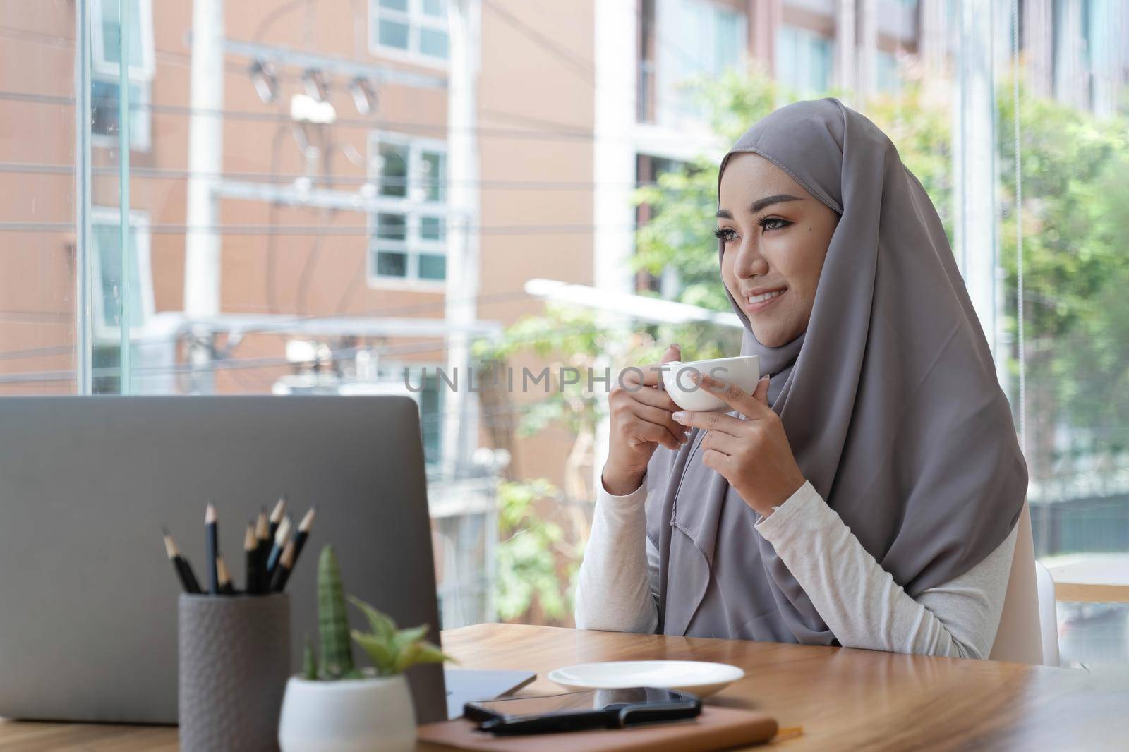 smiling beautiful muslim business woman drinking hot coffee sitting in workspace and looking at window outside thinking work plan..
