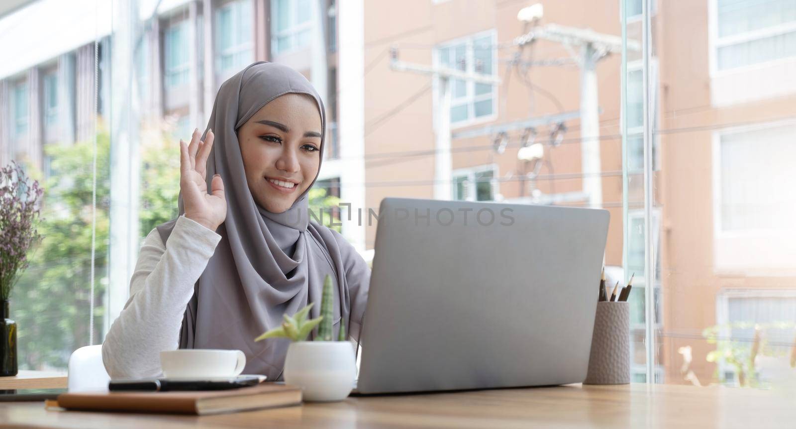 smiling beautiful muslim businesswoman using a laptop for online video meeting sitting at the desk in modern office space, looks at the webcam and waving hello.