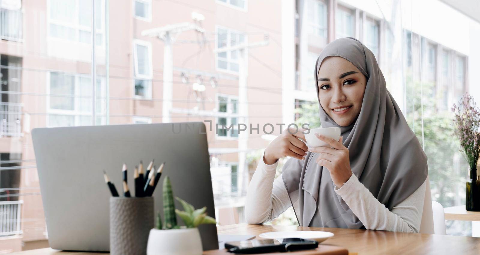 Attractive and cheerful asian muslim female worker or female college student with hijab remote working at the coffee shop, holding a coffee cup..