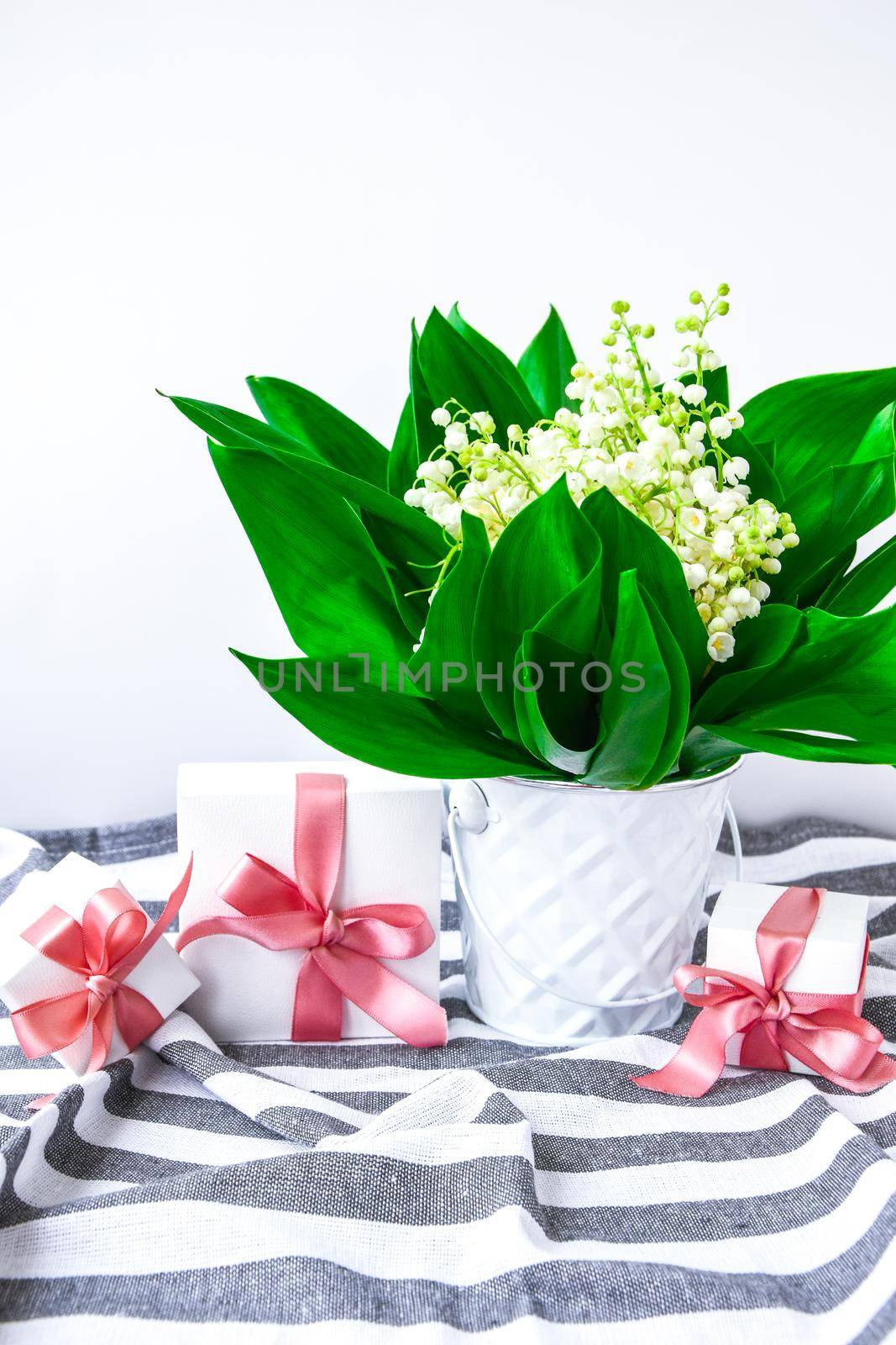 bouquet of lilies of the valley in white bucket with present boxes and pink ribbon on a white table with copy space. concept of holiday background, springtime flowers at home