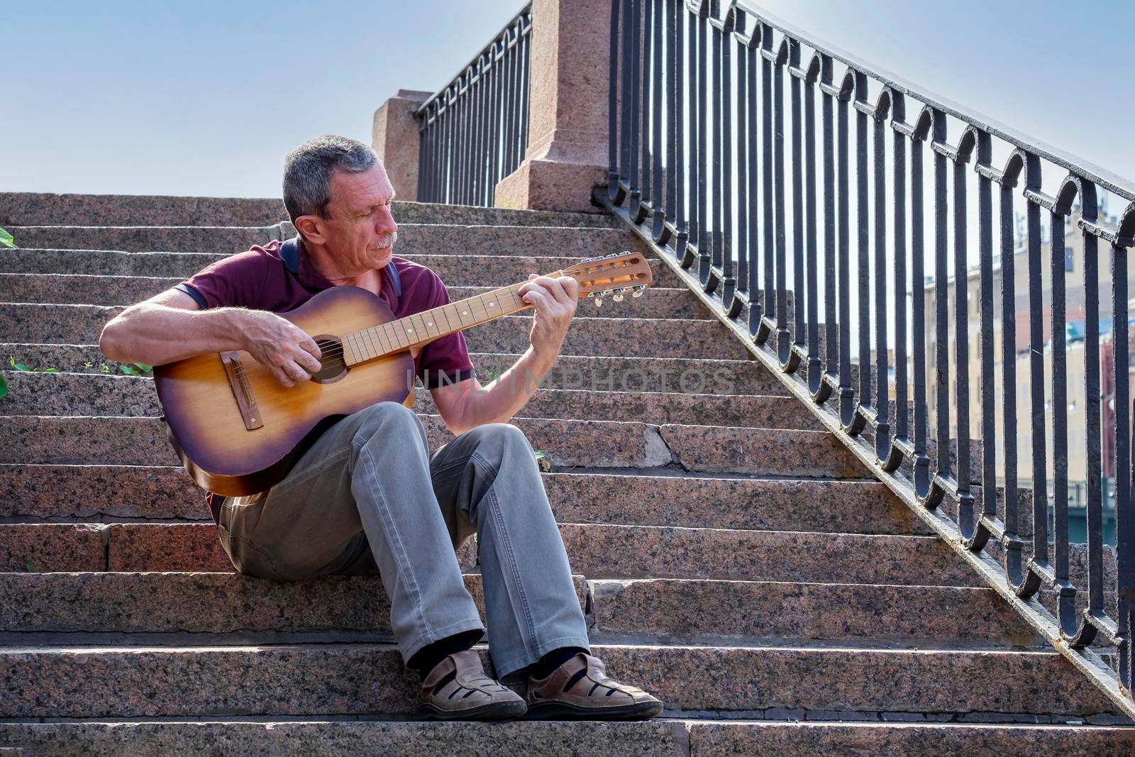 An adult elderly man of retirement age plays an old six-string classical acoustic guitar outdoors while sitting on a granite staircase on a summer evening in the city. Selective focus.