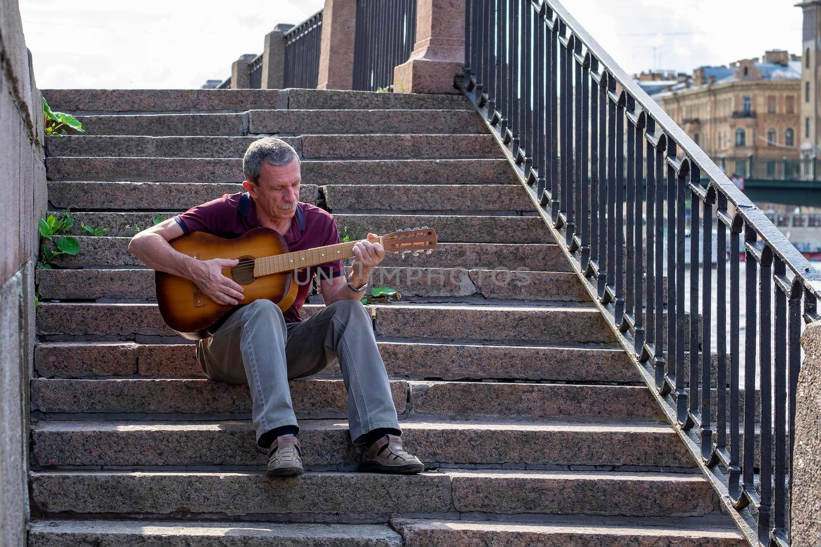 An adult elderly man of retirement age plays an old six-string classical acoustic guitar outdoors while sitting on a granite staircase on a summer evening in the city. Selective focus.