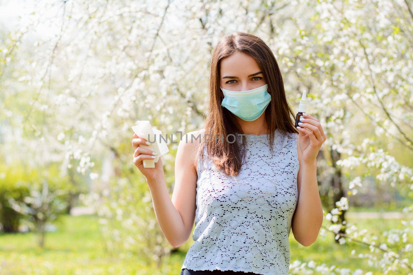 Angry female having allergy standing in blooming spring garden wearing medical mask, holding handkerchief and nasal spray for allergy