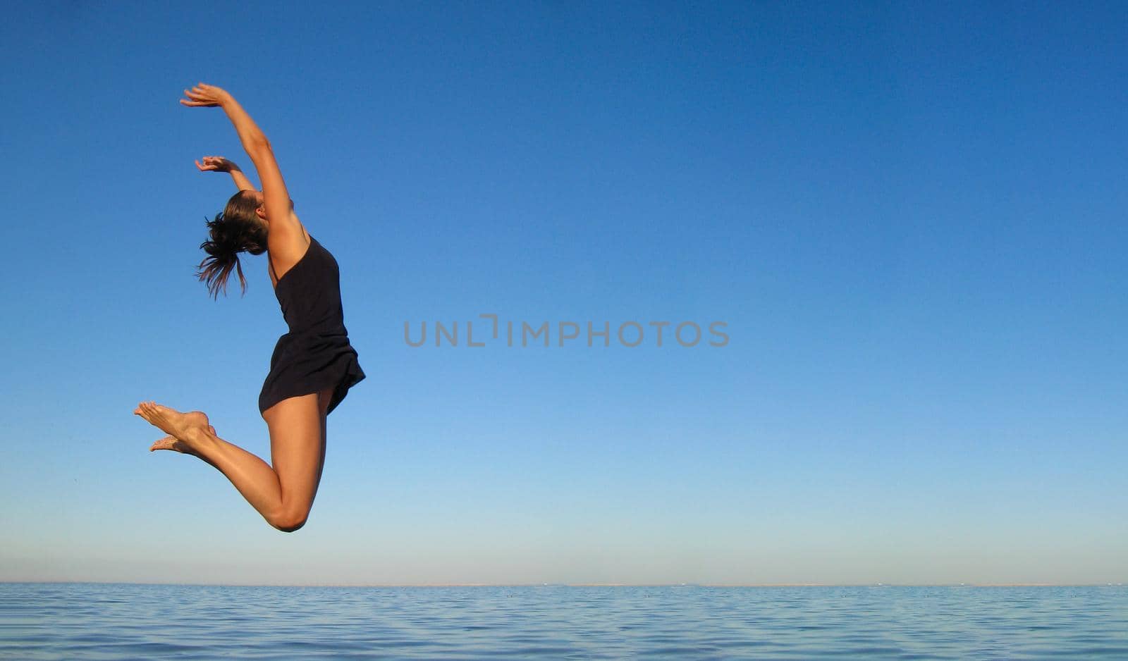 Concept of happiness, freedom, vacation, relaxation. Young girl jumps high in air over sea against sky in dark blue short dress. Banner, copy space