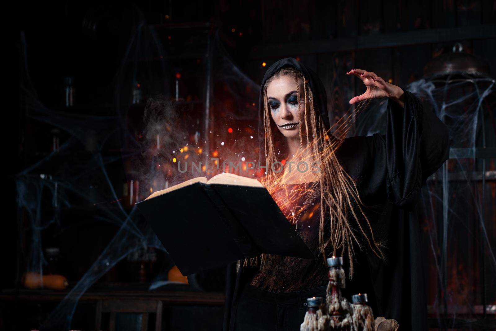 Halloween concept. Witch dressed black hood standing dark dungeon room use magic book conjuring magic spell. Fire sparkles magic Female necromancer wizard gothic interior