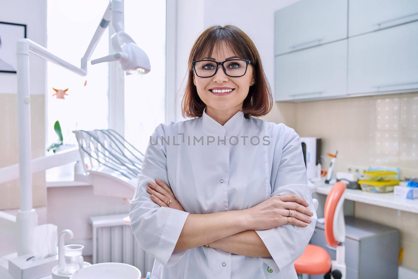 Portrait of smiling mature female dentist looking at camera in office. Confident doctor with crossed arms in clinic. Dentistry, medicine, health care, profession, stomatology concept