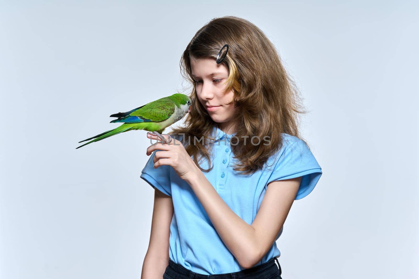 Smiling child girl with green quaker parrot looking at pet on light studio background. Animals, bird owner, childhood, child and pet friendship concept