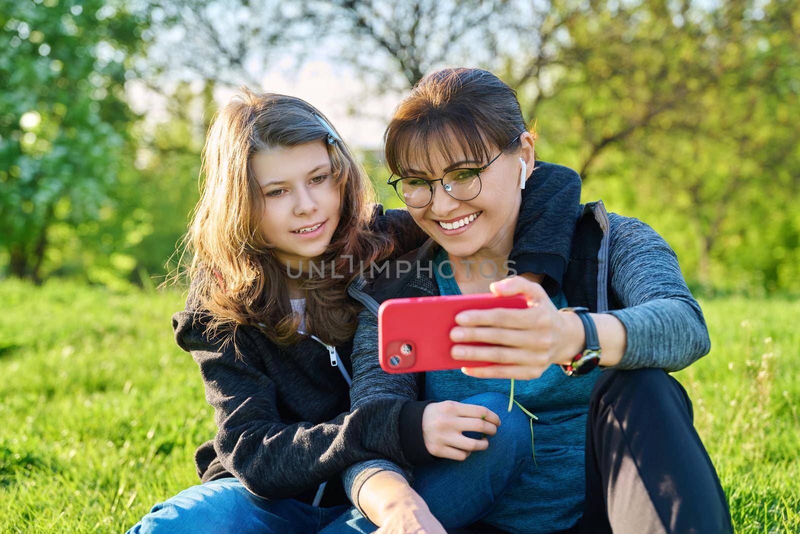 Mom and daughter 11, 12 years old having fun looking together at smartphone screen, outdoor, sitting embracing on grass. Family, leisure, lifestyle, relationship love, mother's day, motherhood concept
