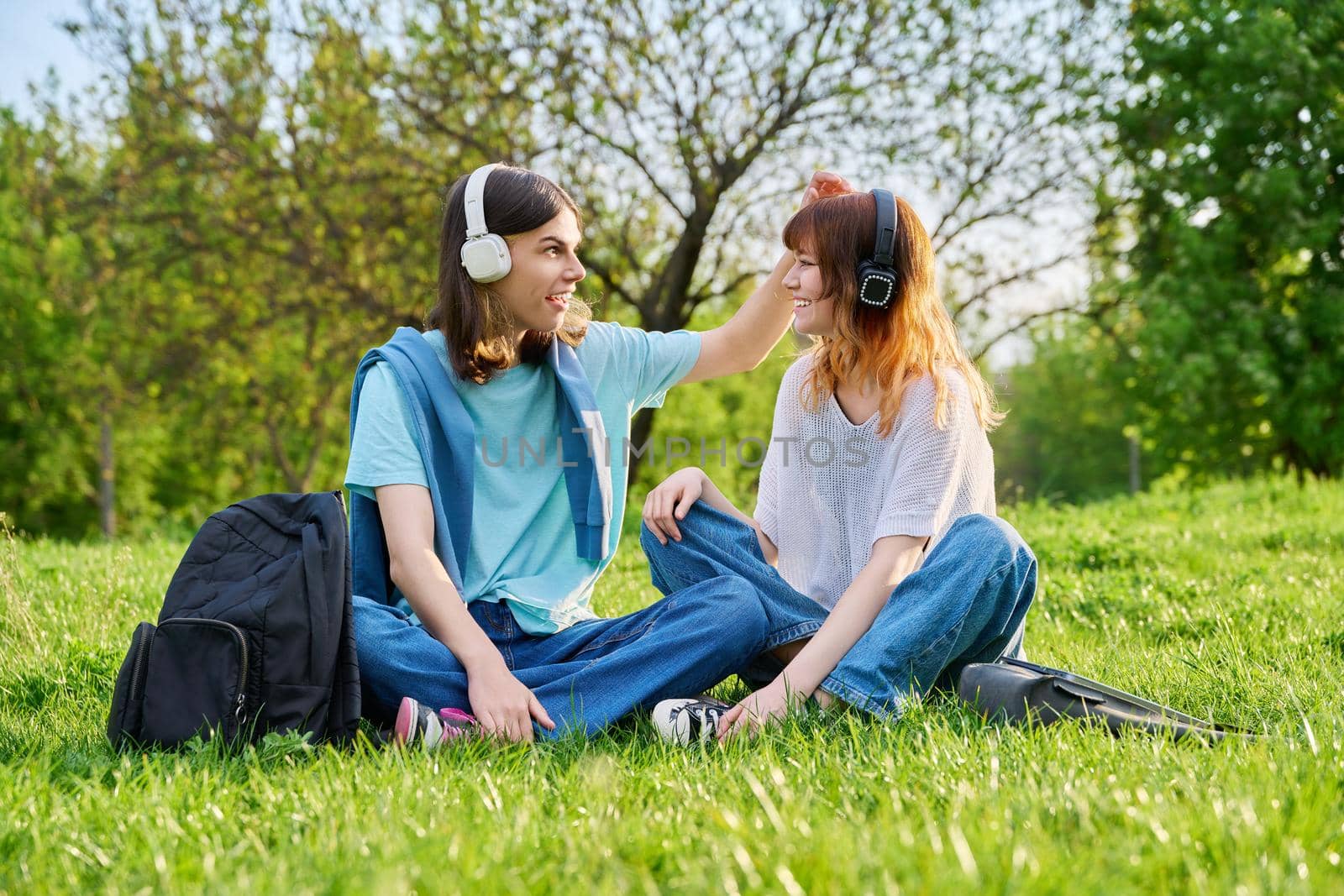 Couple of friends guy and girl 17, 18 years old sitting on grass. Students in headphones with backpacks sitting on campus lawn, talking and laughing. Teens, youth, friendship, education, young people