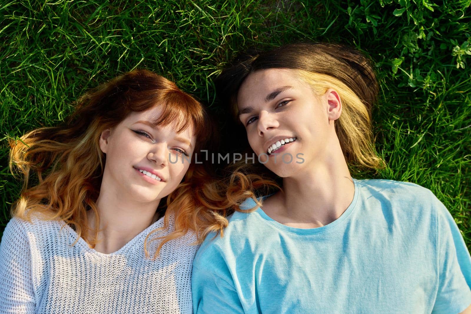 Couple of teenagers friends looking at camera on green grass background. Happy guy and girl 17, 18 years old lying on lawn, top view. Youth, friendship, lifestyle, young people concept