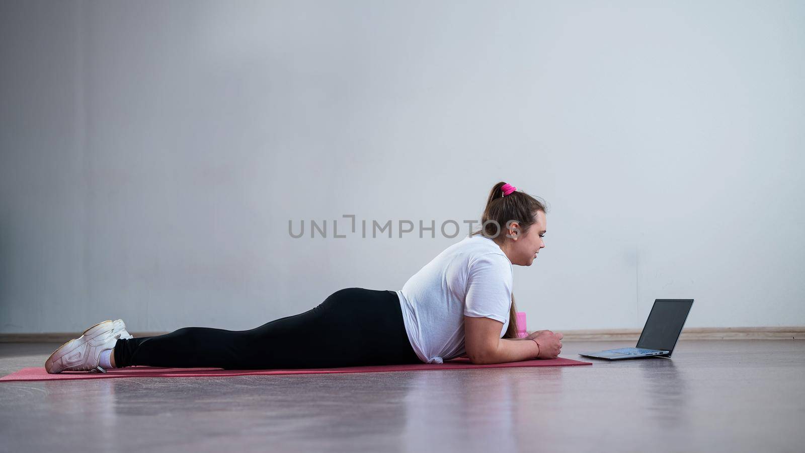 Young Plus Size Woman Stretching At Home Online. Flexible girl practices yoga and watches an online course on a laptop.