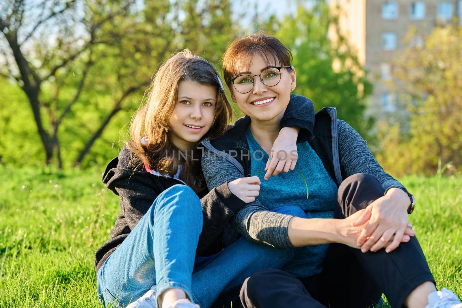Happy mother and daughter hugging sitting on grass, looking at camera. Mom and girl resting together on lawn. Family, happiness, leisure, lifestyle, relationship love, mother's day, motherhood concept