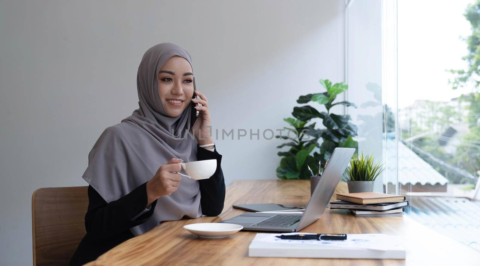 happy beautiful muslim female worker holding hot coffee mug sitting in office and using mobile cell phone viewing online news relaxing. by wichayada