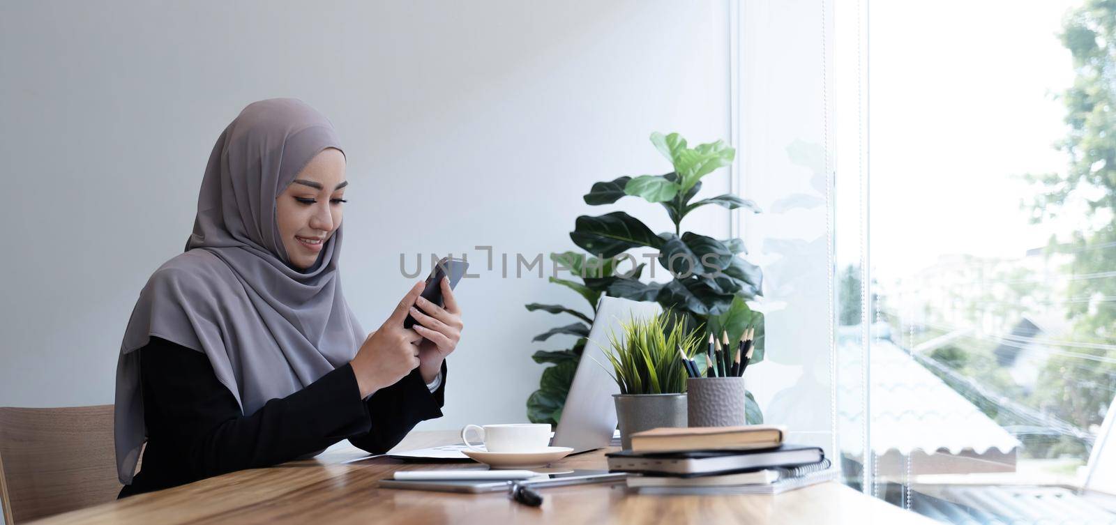 Positive beautiful young muslim woman in purple headscarf using brand new mobile phone, looking at copy space and smiling, using newest mobile application for business, home interior by wichayada