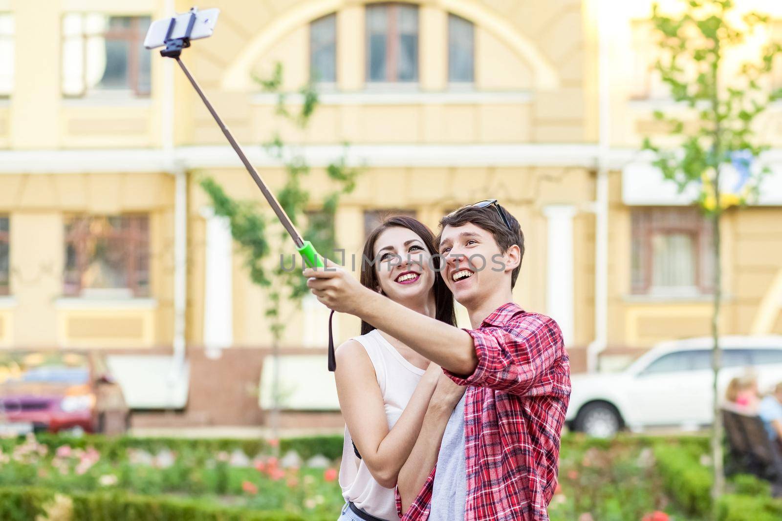 happy young tourists couple taking a selfie with smartphone on the monopod in city. The man is holding the stick and shooting looking at phone with happiness..